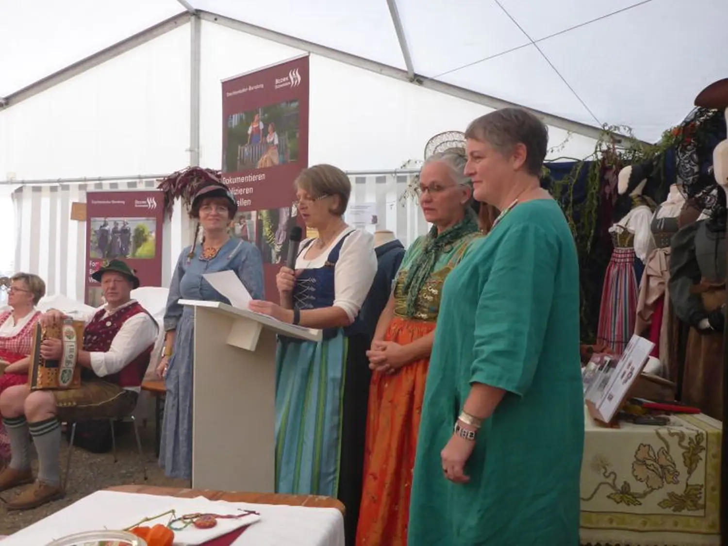 Elbischart – Die Knopfmacherin – Frauen in traditionellen deutschen Dirndln stehen auf einem Podium unter einem Zelt, während ein Mann in Lederhosen in der Nähe Akkordeon spielt.